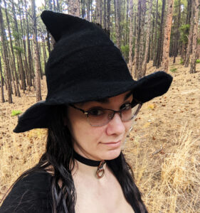 Picture of Jennifer Blackstream standing in a forest wearing a witch hat. She doesn't photograph well, but we love her anyway.